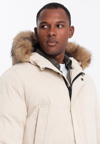 Světlý béžový péřový kabát s kapucí podšitou kožešinou, předním zipem a bočními kapsami. Hladký povrch látky se subtilními detaily šití.