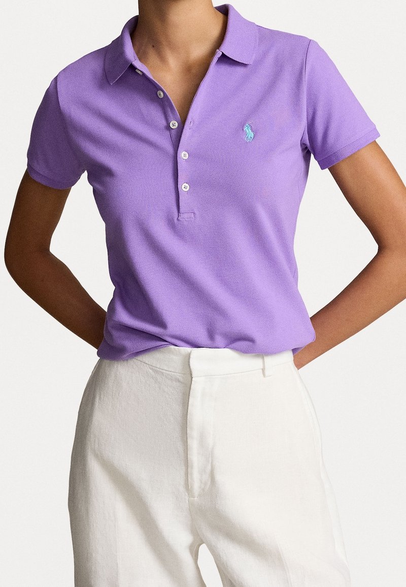 Kvinna iförd en figurnära lila pikétröja med krage och knappar, instoppar i högmidjade vita byxor, med händerna vilande på höfterna.