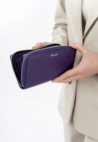 Purple leather wallet with a textured finish, featuring a silver zipper and a logo on the front. Held in hands against a neutral background.