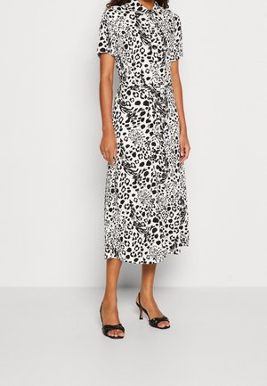 Frau in einem weißen Midikleid mit schwarzem abstraktem Leopardenmuster, kurzen Ärmeln und einem Taillengürtel, kombiniert mit schwarzen offenen Absatzschuhen.