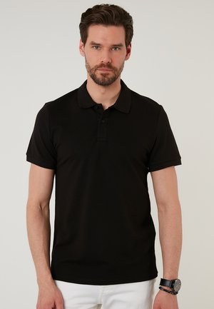 Crna polo majica s kratkim rukavima, teksturirana tkanina i placket s tri dugmeta. Nosena s bijelim hlačama i ručnim satom.