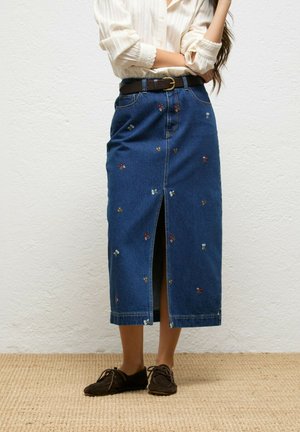 Mujer con una falda midi de mezclilla azul con bordado floral, abertura frontal, cinturón marrón, blusa crema y zapatos marrones con cordones, de pie contra una pared blanca.