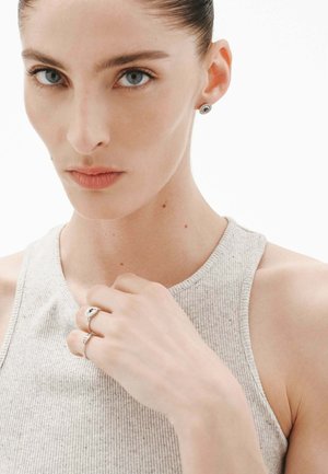 Pendientes de plata en forma de stud y un anillo delgado de plata en una modelo. La modelo lleva una blusa sin mangas de color gris claro y con un escote redondo.