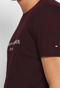 Burgundin värinen puuvillapaita, jossa on pyöreä kaula-aukko. Paidassa on valkoista brodeerattua tekstiä "TOMMY HILFIGER EST. 1985" ja pieni graafinen kuva hihassa.