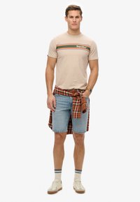 Camiseta de algodón beige con una raya multicolor y el logo de "SuperDry", combinada con pantalones cortos de mezclilla azul y una camisa de cuadros atada a la cintura.