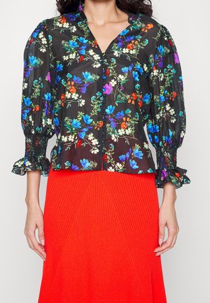 Blouse noire transparente avec imprimé floral vibrant en bleu, violet, rouge et blanc, manches bouffantes, boutonnée à l'avant, associée à une jupe côtelée rouge vif.