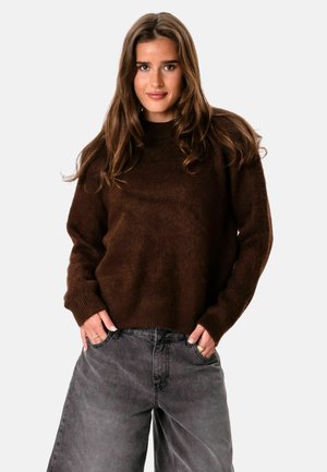 Jeune femme aux longs cheveux bruns portant un pull marron foncé et un jean noir délavé et ample, les mains dans les poches.