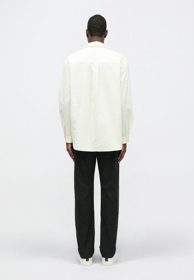 Kremowa bawełniana koszula o prostym kroju, z długimi rękawami i zakładką z tyłu, w zestawieniu z ciemnymi spodniami oraz białymi butami z zielonymi akcentami.