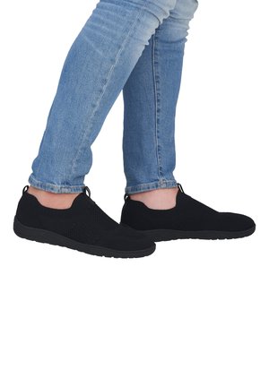 Pieds portant des chaussures noires en mesh sans lacets, associées à un jean slim bleu clair sur fond blanc.