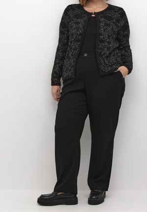 Femme portant un cardigan noir moucheté, un top noir, un pantalon noir taille haute avec bouton et des mocassins à plateforme noirs, debout sur fond blanc.
