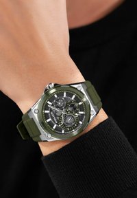 Reloj con correa de goma verde y caja plateada y verde, que presenta un dial negro texturizado y tres subdiales. Muestra marcadores de hora audaces.