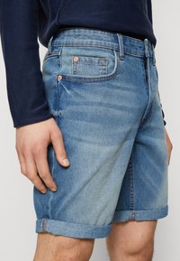 Vaaleansiniset farkkushortsit, joissa on haalistunut pesu, käännetyt lahkeet ja kuparinväriset yksityiskohdat. Mukana sivu- ja takataskut.