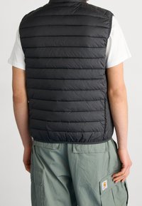 Czarna, pikowana kamizelka z poziomym przeszyciem, wykonana z lekkiego materiału. Posiada kołnierz i prosty dół, noszona na białej koszuli.