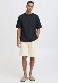 T-shirt en coton noir, coupe décontractée, manches courtes. Short beige clair jusqu'aux genoux avec un motif à carreaux subtil. Chaussures grises à enfiler.