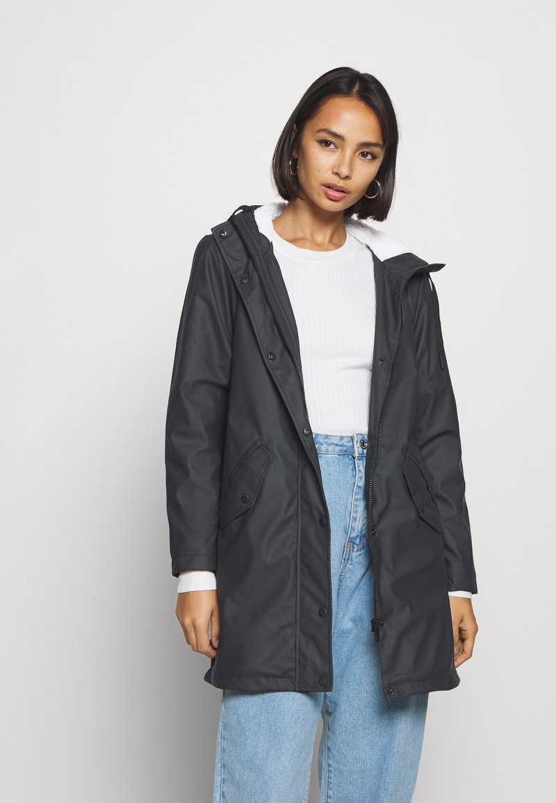 Svart parka-jakke med en myk hvit krage, knappe- og glidelåslukking, sidelommer og en avslappet passform, kombinert med lyseblå jeans.