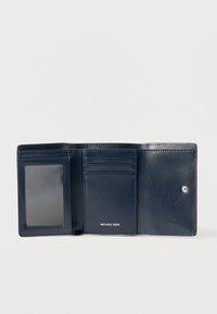 Tamnoplavi kožni novčanik ima više pretinaca za kartice, prozirni džep za osobnu iskaznicu i zatvarač na kopču. Naziv marke "MICHAEL KORS" utisnut je iznutra.