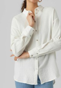 White button-up shirt made of textured fabric, featuring a collar, long sleeves, and pearl-like buttons. Casual fit with a slightly curved hem.