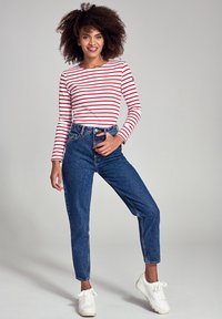 Chemise à rayures rouges et blanches à manches longues, jeans bleus taille haute, baskets blanches. Le mannequin se tient avec une main sur la hanche, souriant.