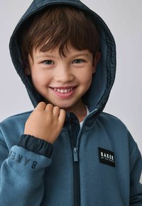 Donkerblauwe fleece hoodie, met een rits aan de voorzijde, elastische manchetten en een zwart "BAKER" logo patch op de borst.