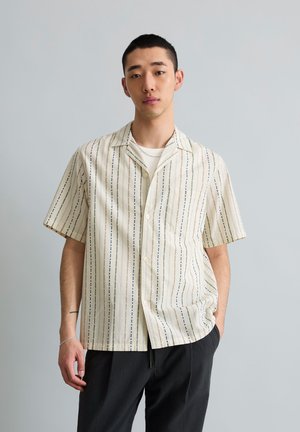 Młody mężczyzna o krótkich włosach nosi kremową koszulę z krótkim rękawem z pionowymi paskami i czarne spodnie, stoi na tle jednolitego szarego tła.