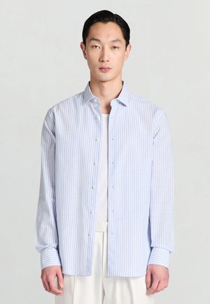 Chemise à rayures bleu clair en coton avec col, manches longues, et boutons blancs, portée par-dessus un débardeur blanc.