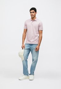 Svetlo roza polo majica s tremi gumbi, združena s svetlo modrimi kavbojkami. Na nogah ima bele superge in v roki svetlo kapo.