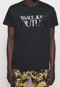 Schwarzes T-Shirt mit glänzendem "Versace Jeans Couture"-Schriftzug, getragen mit schwarzen Hosen, die ein gold- und weißes Barockmuster sowie wiederholte Markenlogos aufweisen.