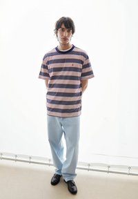 Gestreept t-shirt in roze en marineblauw, korte mouwen, ronde hals, met een klein rood accent. Lichtblauwe spijkerbroek en zwarte schoenen. Neutrale achtergrond.