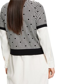 Suéter de punto recortado en blanco y negro con un patrón cuadrado, dobladillo y puños de canalé negros, llevado sobre una camisa blanca con cuello.