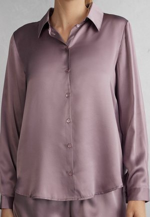 Mujer vistiendo una camisa satinada de manga larga color malva con botones y cuello, mostrada desde los hombros hasta las caderas sobre un fondo liso.