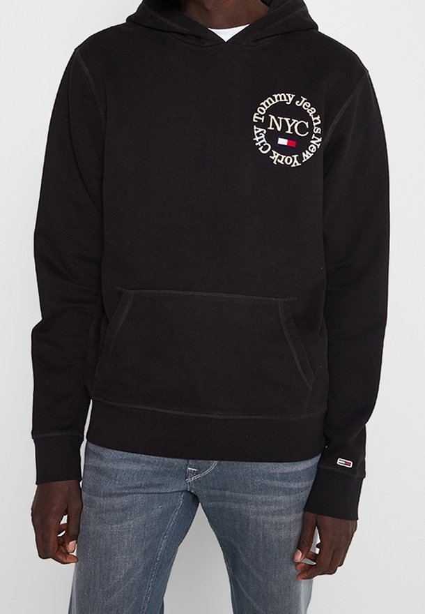 Czarna bluza z kapturem z napisem "Tommy Jeans New York City NYC" na piersi oraz małą flagą Tommy na mankiecie rękawa, noszona z jeansami w średnim praniu.