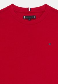Camiseta de algodón roja con cuello redondo, que presenta un pequeño logo azul, blanco y rojo en el lado izquierdo del pecho y detalles a rayas en el cuello.