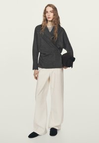 Czarna szara bluzka z długimi rękawami, zestawiona z szerokimi białymi spodniami. Osoba trzyma brązowy przedmiot i ma na sobie czarne buty typu slip-on.