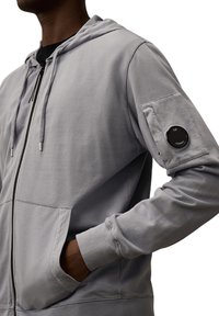 Lichtgrijze zip-up hoodie van zachte stof, met een vozak pocket, verstelbare capuchon en een rond logo-patch op de linkermouw.