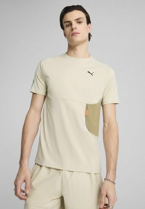 Beige kortærmet t-shirt med rund halsudskæring, der har en lynlåslomme i en mørkere nuance, samt et sort logo på brystet.