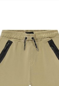 Beige joggers met een elastische tailleband, zwarte ritszakken aan de zijkanten en een contrasterend trekkoord. Gladde textuur met subtiele stikdetails.