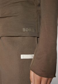Ruskea pitkähihainen paita ja pehmeäkangas-housut, joissa on vaalealla värillä "BORG"-logo kummassakin tuotteessa. Saumadetailit näkyvissä.