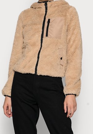 Beige fleecejack met rits en twee beige stoffen zakken, gedragen met zwarte broek, toont bovenlichaam en ontspannen armen langs het lichaam.