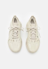 Ett par krämfärgade, snörade sportskor med nätmaterial och gummiyttersulor, visade ovanifrån på en vit bakgrund.