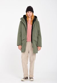 Parka verte avec capuche, fermetures éclair et à pression, deux poches avant, portée par-dessus un hoodie noir et une chemise rose, associée à un pantalon en velours côtelé clair.