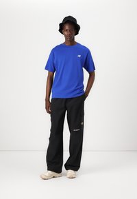 Blauwe T-shirt met korte mouwen en logo, gecombineerd met zwarte cargobroek met zijzakken en een gouden accent. Beigekleurige sneakers en een zwarte bucket hat.