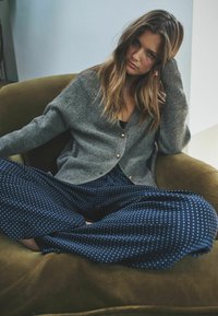 Grå strikket cardigan med gyldne knapper, båret over en sort bluse, parret med blå polkaprikkede vidde bukser, siddende på en grøn stol.