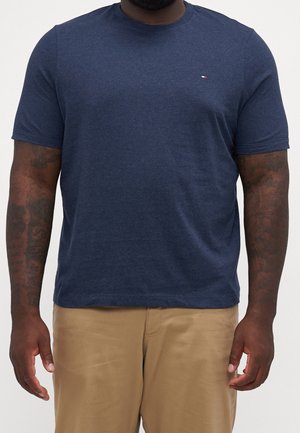 Mann trägt ein marineblaues kurzärmeliges T-Shirt mit kleinem Logo auf der Brust und beige Hosen, steht mit entspannt an den Seiten hängenden Armen.