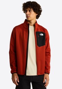 Rode softshell jas met een zwarte borstzak met ritssluiting en zwarte accenten. Voorzien van een hoge kraag en elastische manchetten. Draagt over een licht shirt.