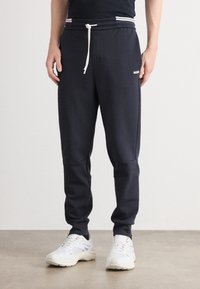 Mand iført navy joggers med hvid snor og hvide sneakers, stående på et ensfarvet gulv mod en lys baggrund.