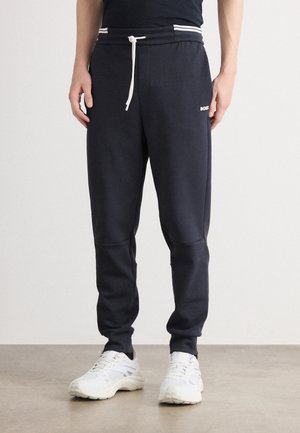 Mann in marineblauen Jogginghosen mit weißem Kordelzug und weißen Sneakern, der auf einem schlichten Boden vor einem hellen Hintergrund steht.