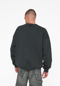 Sweatshirt gris foncé avec un col rond et des épaules tombantes. Présente une broderie texturée au dos et des poignets côtelés.