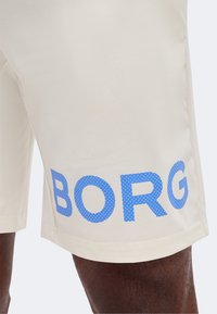 Μπεζ σορτς με μεγάλα μπλε κουκκίδια που σχηματίζουν το όνομα "BORG" στο αριστερό μηρό, που φοριέται από άτομο με σκουρόχρωμο δέρμα.