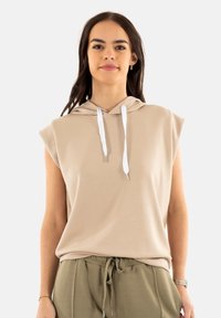 Sudadera sin mangas en beige con cordón, fabricada en tejido suave y con un corte holgado. Combinada con pantalones sueltos en verde oliva.