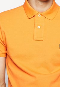 Orange polo t-shirt i ett texturerat tyg, med en klassisk krage, två knappar och en liten broderad logotyp på vänster bröst.